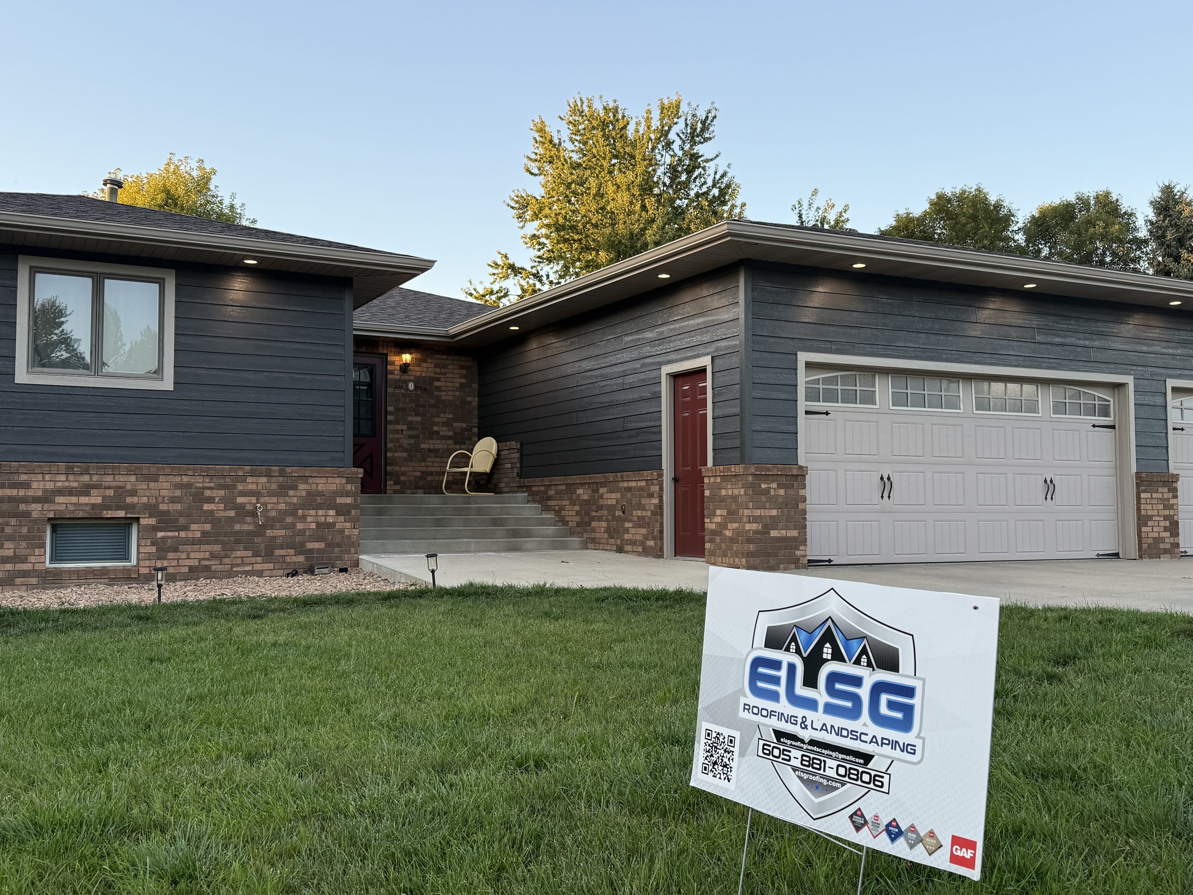 Completed charcoal siding on a brick-base split-level, with the ELSG yard sign standing in the foreground lawn: logo, phone number and the GAF badge strip readable on the sign face.