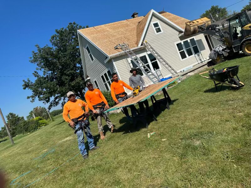 Four crew in orange hi-vis walking a plywood cut table in front of a farmhouse stripped to bare OSB decking, with a skid loader and a debris bucket behind them.