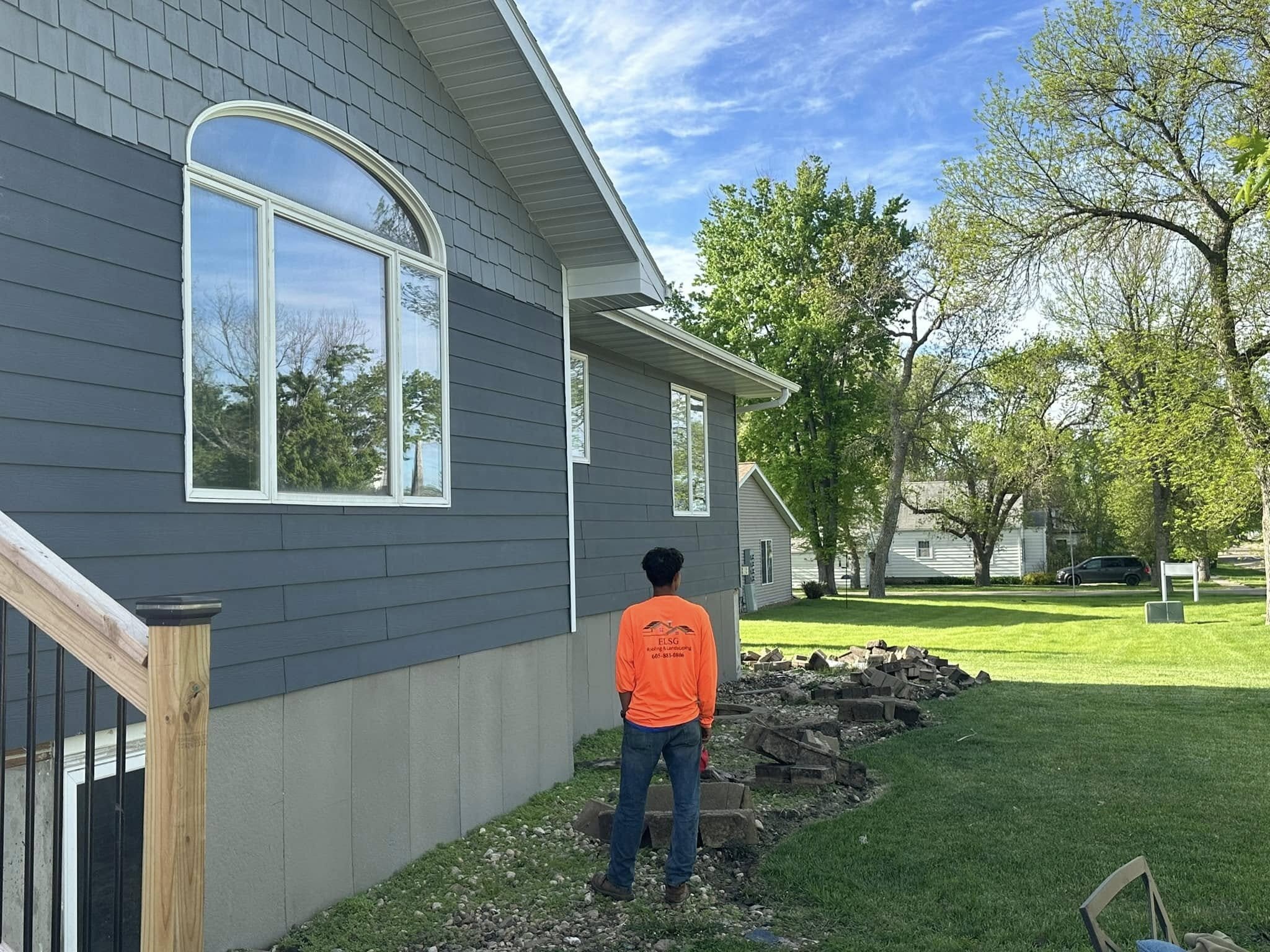 A worker in an ELSG-branded orange shirt, the logo and phone number readable on the back, standing beside a gray-sided house with an arched window, with retaining-wall block staged across the lawn.