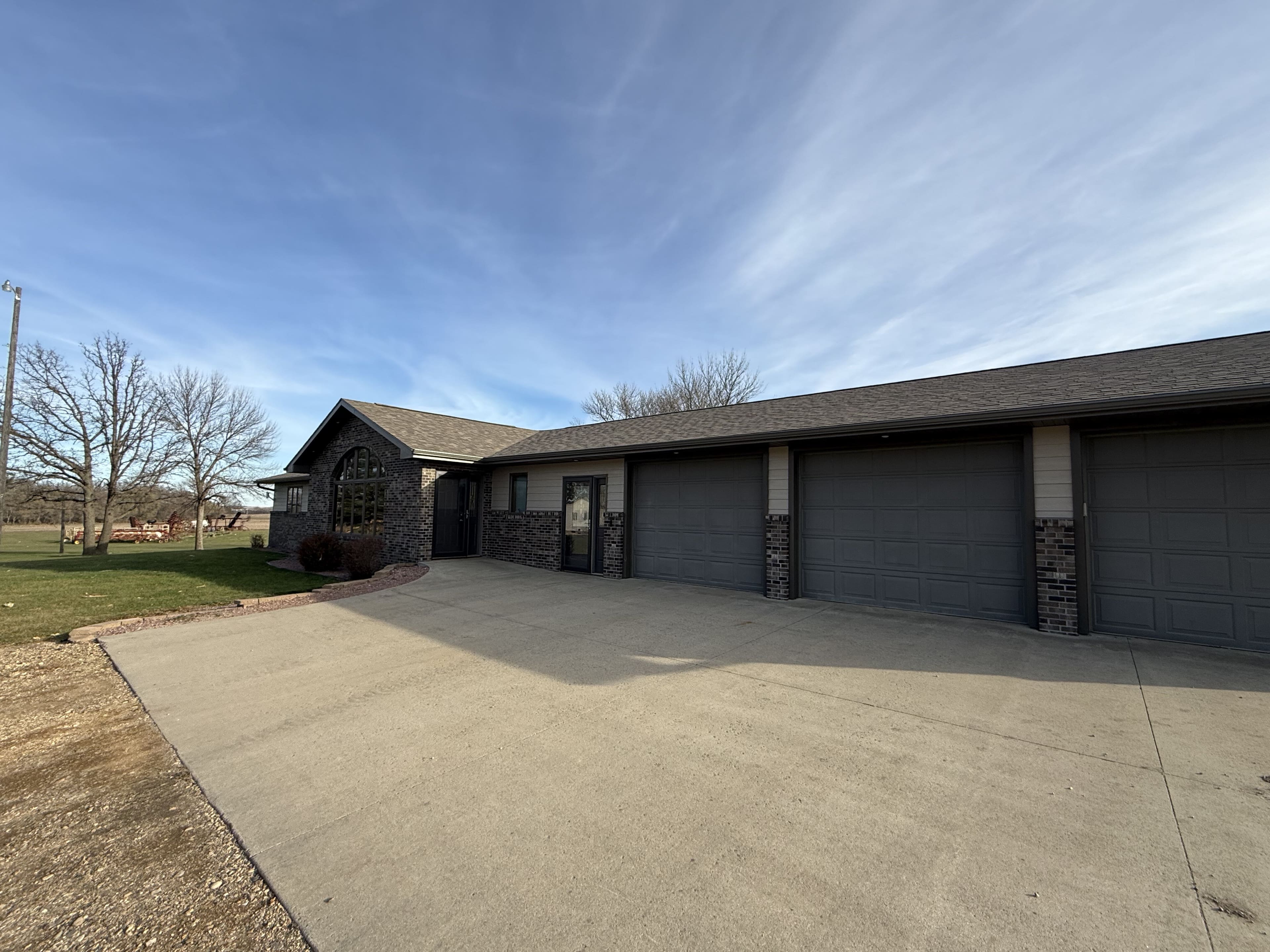 A single-story brick ranch home with new architectural shingles running the length of the roof, a row of garage stalls and a wide concrete drive under a clear sky