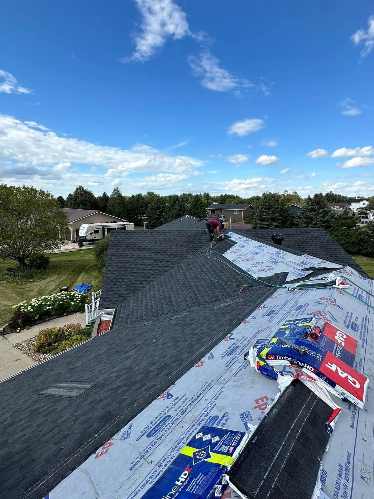 Charcoal shingles mostly laid across the main field, a worker nailing at the ridge, and GAF Timberline HDZ bundles staged on Epilay underlayment on the right slope with their wrappers legible, hydrangeas and a white porch rail below