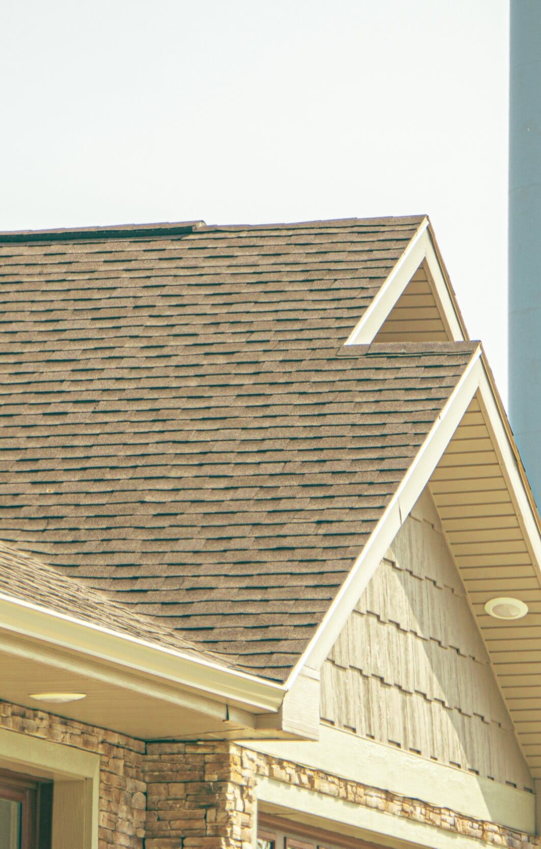 A tan shingle roof meeting a cream shake-panel gable above a stone column
