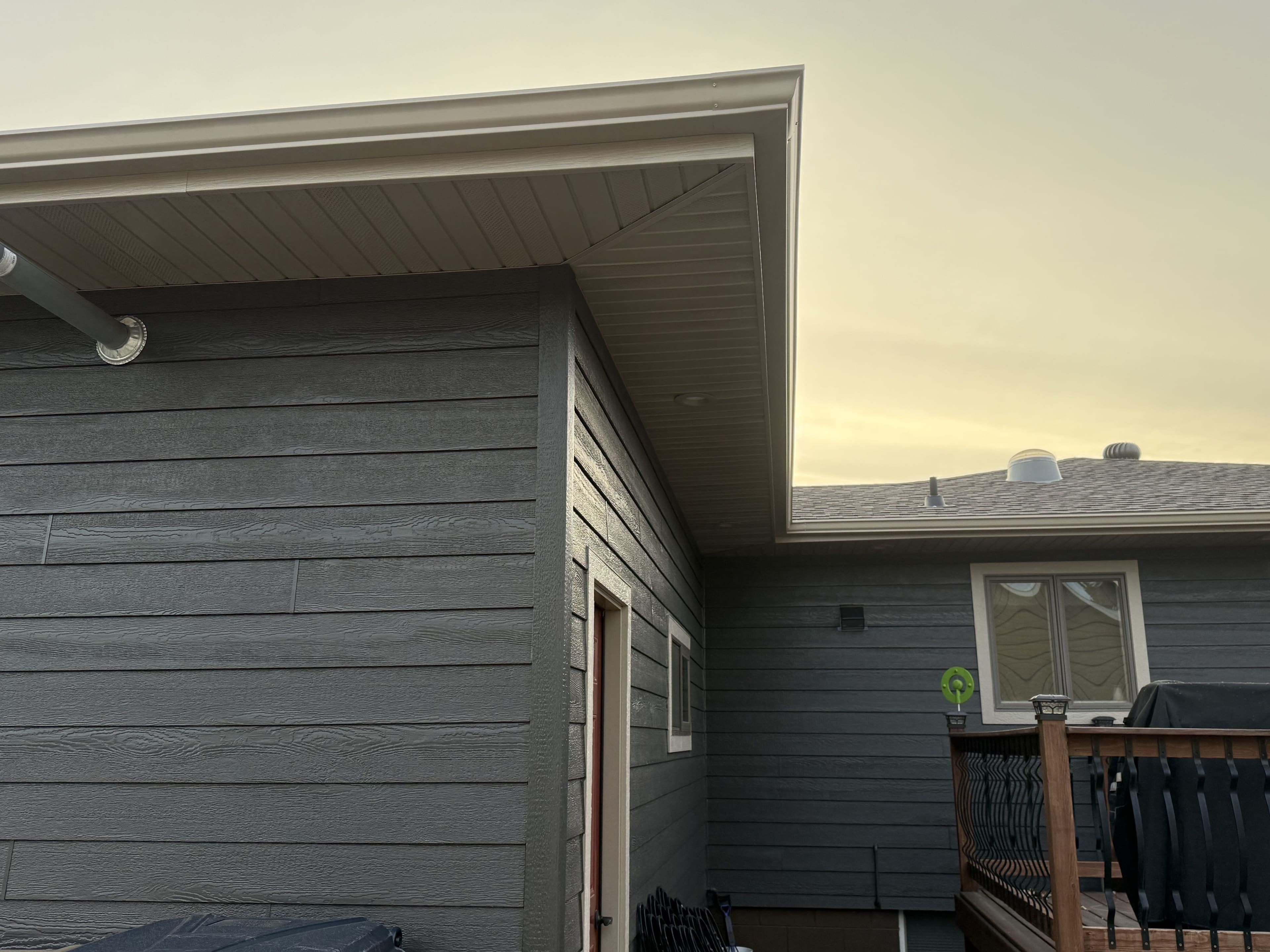 Charcoal engineered-wood lap siding meeting white soffit and fascia at a gable corner, with the downspout running down and a deck rail below