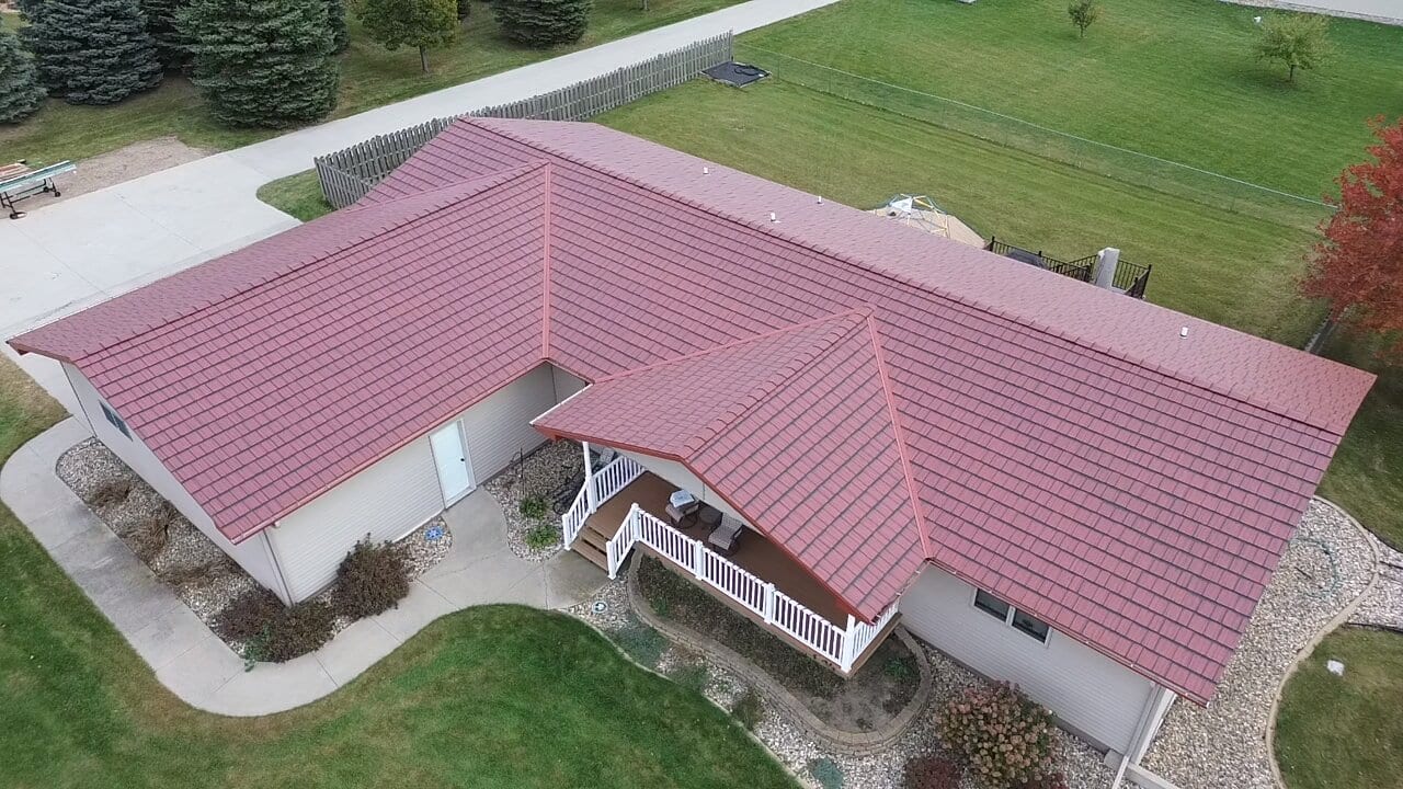 A drone view straight down onto a finished burgundy steel-shingle roof on a ranch home, with a white deck rail and concrete drive below