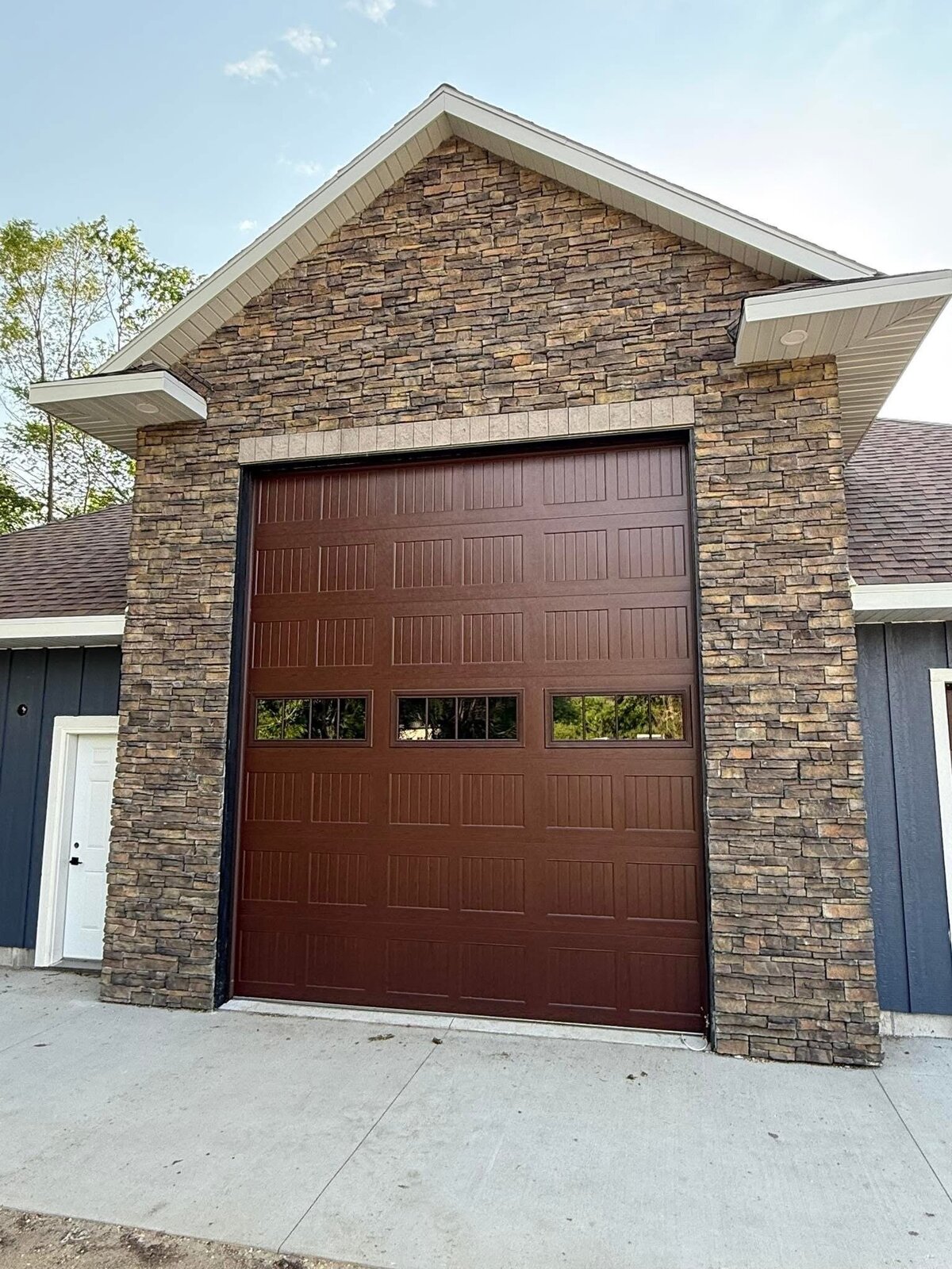 After. The finished garage elevation straight on: full stacked-stone gable and piers, cream soffit and fascia, dark red-brown carriage garage door with a three-lite window row, navy board-and-batten either side, white service door at the left, poured concrete apron, green trees