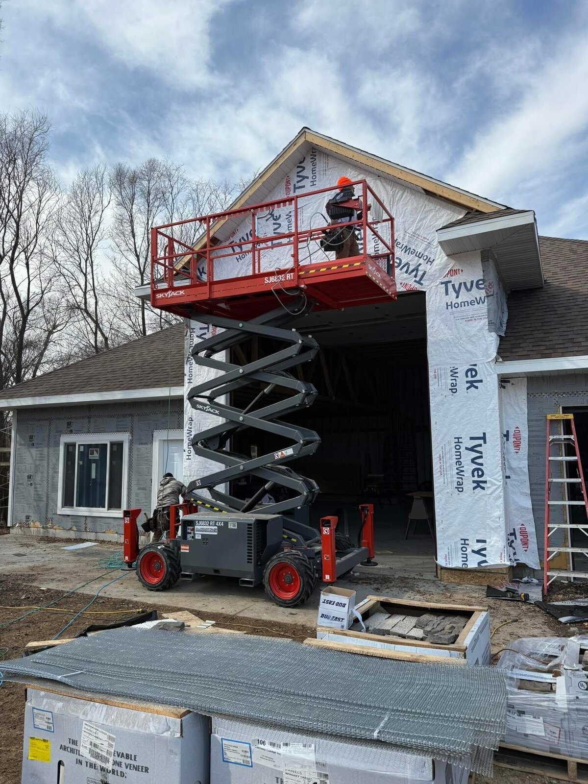 Before. The garage gable wrapped in DuPont Tyvek HomeWrap over the open bay, a worker on the raised Skyjack platform working the wrap, a second worker at the machine below, stone-veneer pallets staged in the foreground, bare trees