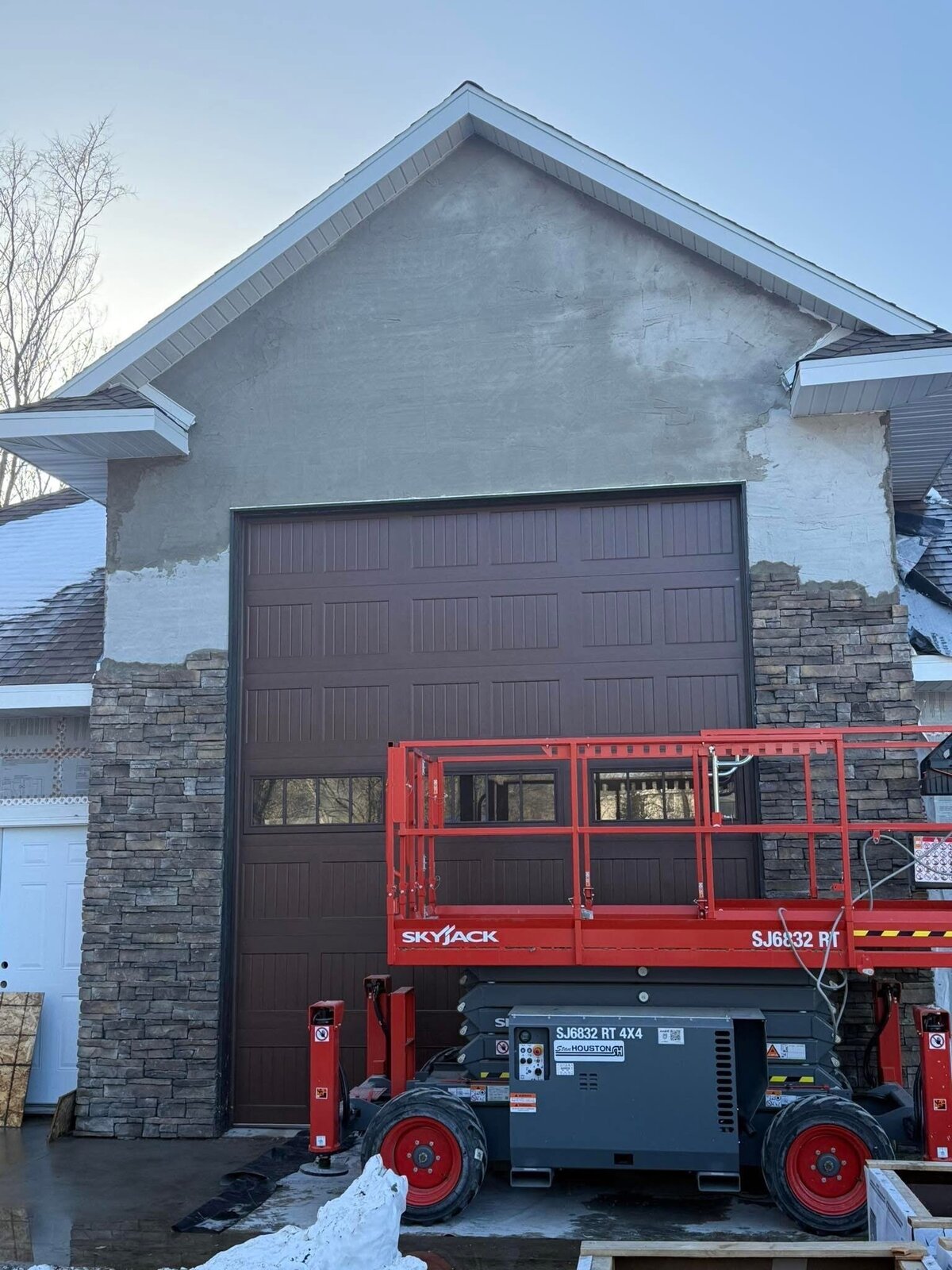 During. The same garage gable with a grey mortar scratch coat across the field and stone veneer run up both piers, ragged at the top where the course stops, garage door now hung, snow on the ground
